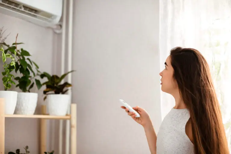 Young woman adjusts the temperature of the air conditioner using the remote control in room with a lot of plants at home
